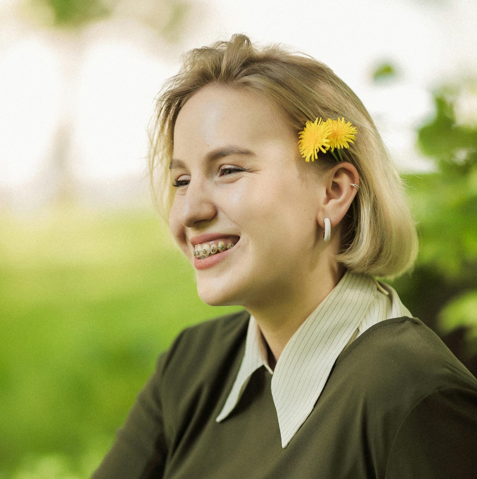 Mujer de cabello rubio, vistiendo un top top verde con cuello blanco. Ella está sonriendo y tiene una flor amarilla en el pelo. En su sonrisa tiene brackets.